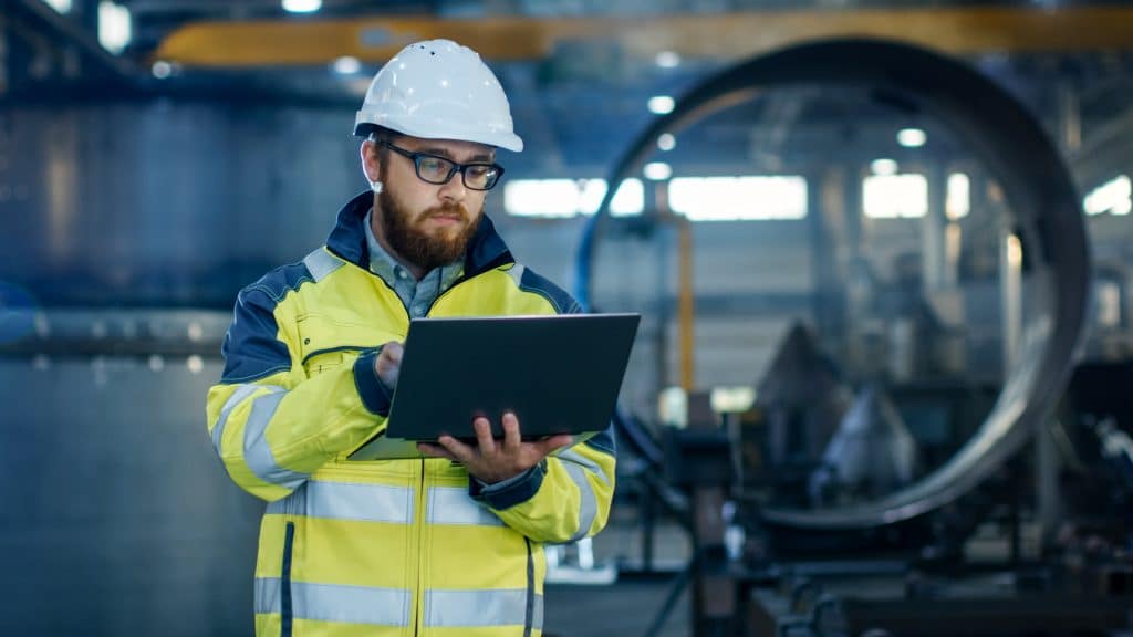 Industrial Engineer in Hard Hat Wearing Safety Jacket Uses Laptop. He Works in the Heavy Industry Manufacturing Factory with Various Metalworking Processes are in Progress.